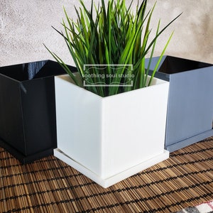 May include: Three square plant pots, one black, one white, and one gray, with a green plant in the white pot. The pots have a square base and are made of a smooth material. The text "soothing soul studio" is visible on the white pot.