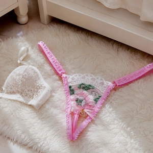 Peut inclure: Un soutien-gorge en dentelle blanche et un string rose et blanc sont présentés sur un tapis blanc duveteux. Le string présente un motif floral avec des bretelles roses et un petit charme doré. Le soutien-gorge a des détails en dentelle délicats.