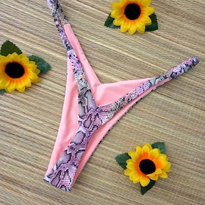 Könnte beinhalten: Ein pinkfarbener Bikini-Slip mit Schlangenmuster-Besatz, vor einem gewebten Hintergrund. Der Bikini ist von drei gelben Sonnenblumen mit grünen Blättern umgeben, was eine sommerliche Atmosphäre schafft.