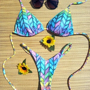 Può includere: Un colorato set bikini con un motivo geometrico nei toni del blu, viola e giallo. Il set comprende un top a triangolo e uno slip a perizoma. Un paio di occhiali da sole e due girasoli sono presenti nell'immagine.