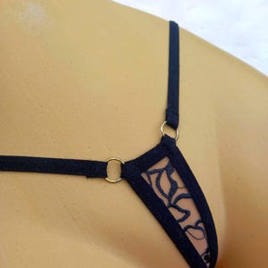 May include: Black lingerie thong with a delicate floral lace design. The thong features thin straps and gold-coloured metal rings at the connection points. The lace is visible against a nude background.