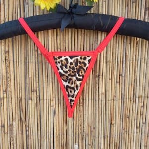 Op de afbeelding: Een rode en luipaardprint string bikini broekje hangt aan een zwarte gewatteerde hanger. De bikinibroek heeft een driehoekige vorm met een luipaardprint middenstuk en rode rand. Gele zonnebloemen en een zwarte strik versieren de bovenkant.