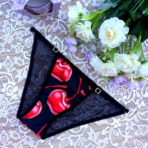 May include: Black lace underwear with a red cherry print. The underwear has gold metal clasps on the sides.