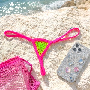 May include: A neon pink thong bikini with a neon green mesh center, a pink mesh bag, and a bedazzled phone case with various gems and rhinestones are displayed on a white rock near the ocean.