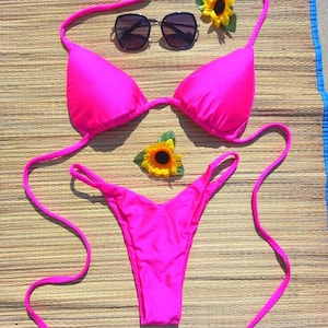 May include: A bright pink bikini set with matching string ties, a triangle top, and a thong bottom. The set is accessorized with dark sunglasses and two yellow sunflowers, all arranged on a woven mat.