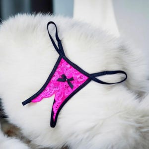 May include: A hot pink lace thong with black trim and straps. A small black bow with a rhinestone center adorns the front. The lingerie is displayed on a white, fluffy surface.