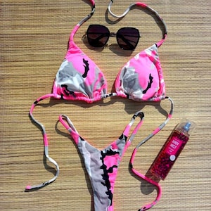May include: A pink, grey, and black camouflage print bikini set with matching string ties. The set includes a triangle top and a thong-style bottom. Black sunglasses and a bottle of body mist are also in the image.