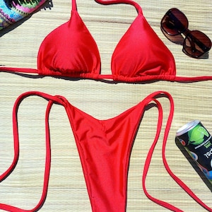 Può includere: Un bikini rosso acceso, composto da un top a triangolo e slip sgambati, disposto su un tappetino intrecciato. L'immagine include occhiali da sole, un fermaglio per capelli e una lattina di bibita. Il costume da bagno è realizzato in tessuto lucido.