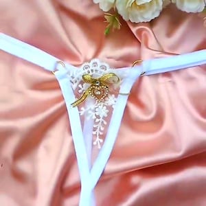 May include: White thong underwear with a decorative gold bow and heart-shaped embellishment. The lingerie features delicate lace detailing and gold ring accents. The background is a soft, peach-colored fabric with white flowers.