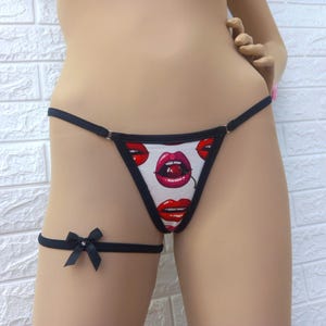 May include: White thong underwear with a pattern of red lips and cherries. The thong has black trim and a matching garter with a black bow. The lingerie is displayed against a white brick wall.