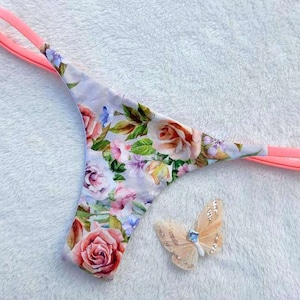 Könnte beinhalten: String-Tanga mit floralem Druck und einem rosa elastischen Bund und Trägern. Der Stoff weist ein Muster aus Rosen in Rosa-, Pfirsich- und Lilatönen mit grünen Blättern auf. Ein dekorativer Schmetterling befindet sich neben der Unterwäsche.