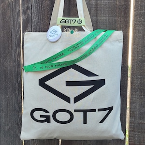 May include: A white canvas tote bag with a black GOT7 logo and green ribbon with the text "IS OUR NAME" on it. The bag also has a green button with the GOT7 logo on it.