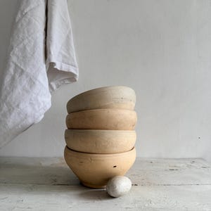 May include: A stack of four light brown wooden bowls with a rounded shape, sitting on a white wooden surface. A small, light-colored stone sits near the bowls. A white linen cloth hangs in the background.