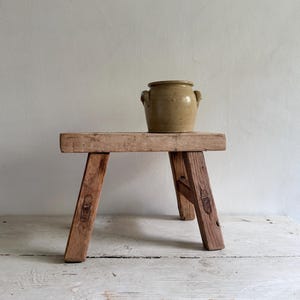 May include: A small, rectangular wooden stool with four angled legs and visible joinery details, featuring a weathered, light brown finish. Resting on the flat top of the stool is a single, cylindrical, tan-coloured ceramic pot with two small handles.