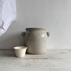 May include: A grey ceramic crock with handles and a small, cream-coloured ceramic cup with holes. The objects are on a weathered, white wooden surface. A white cloth hangs in the background.