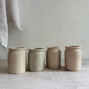May include: A set of four cylindrical ceramic crocks in varying shades of cream, beige, and light grey. Each vessel features a smooth, glazed finish, a slightly flared rim, and a simple, sturdy base, presented in a row for storage or display.
