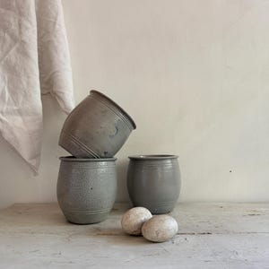 May include: Three grey stoneware crocks with a smooth, glazed finish and a cylindrical shape. Two crocks sit upright on the surface, while the third is tilted at an angle on top of one. Two small, textured, off-white oval stones rest in front.