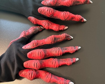 May include: A pair of black gloves featuring long, textured red finger extensions that resemble creature claws. The fingers have a wrinkled, organic surface with dark shading and pointed, off-white tips, extending from a smooth black fabric base.