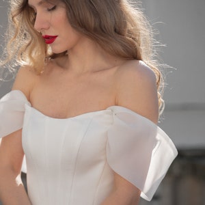 May include: A woman wearing a white off-the-shoulder wedding dress with a sweetheart neckline and a fitted bodice. The dress has a tulle skirt and sheer, puffed sleeves.
