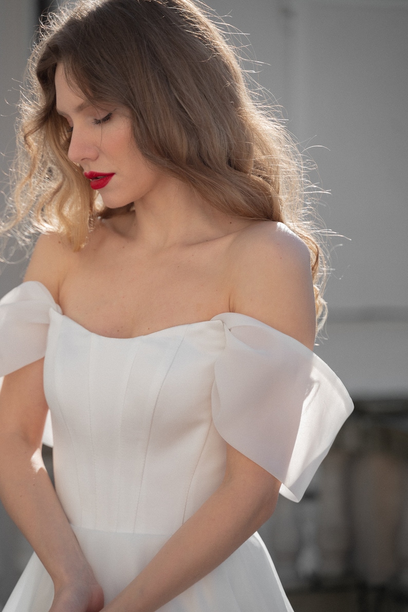 May include: A woman wearing a white off-the-shoulder wedding dress with a sweetheart neckline and a fitted bodice. The dress has a tulle skirt and sheer, puffed sleeves.