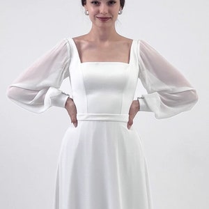 Puede incluir: Vestido de novia blanco con escote cuadrado y mangas largas transparentes. El vestido presenta un corpi&ntilde;o ajustado y una falda fluida. Un cintur&oacute;n fino ci&ntilde;e la cintura. La modelo lleva una tiara y pendientes.