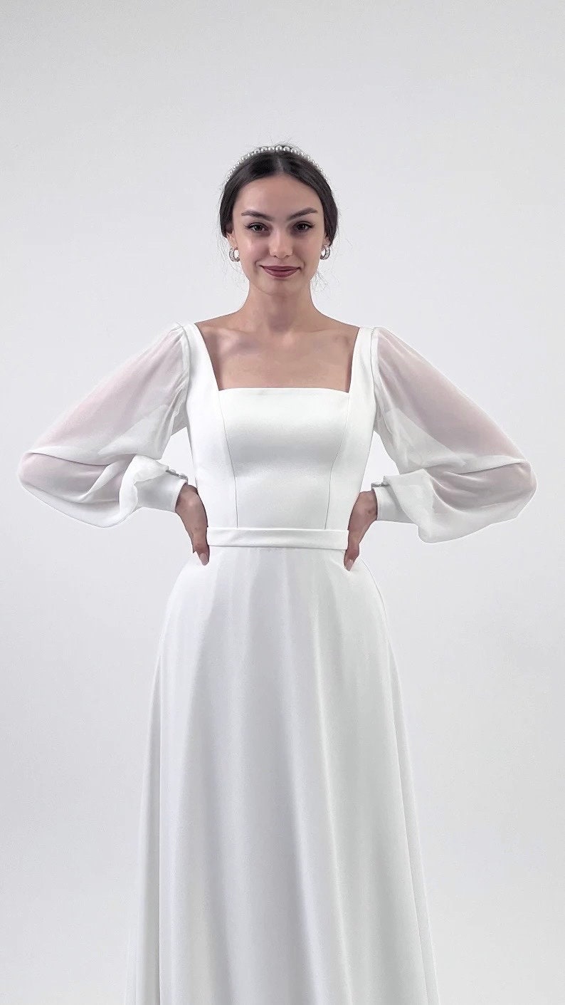 Puede incluir: Vestido de novia blanco con escote cuadrado y mangas largas transparentes. El vestido presenta un corpi&ntilde;o ajustado y una falda fluida. Un cintur&oacute;n fino ci&ntilde;e la cintura. La modelo lleva una tiara y pendientes.