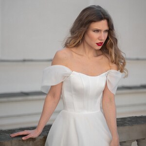 May include: A woman wearing a white off-the-shoulder wedding dress with a fitted bodice and a flowing skirt. She is standing in front of a white wall and has her hair pulled back.