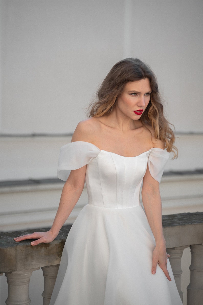 May include: A woman wearing a white off-the-shoulder wedding dress with a fitted bodice and a flowing skirt. She is standing in front of a white wall and has her hair pulled back.