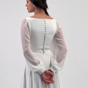 Puede incluir: Vestido de novia blanco con mangas largas y transparentes y escote cuadrado. La espalda presenta una fila de peque&ntilde;os botones. El vestido tiene un corpi&ntilde;o ajustado y una falda fluida. Se lleva una diadema de perlas.