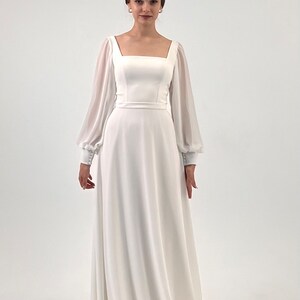Puede incluir: Un vestido de novia blanco con escote cuadrado y mangas largas transparentes. El vestido presenta un corpi&ntilde;o ajustado y una falda fluida. Las mangas tienen pu&ntilde;os abotonados. La modelo lleva una tiara y pendientes.