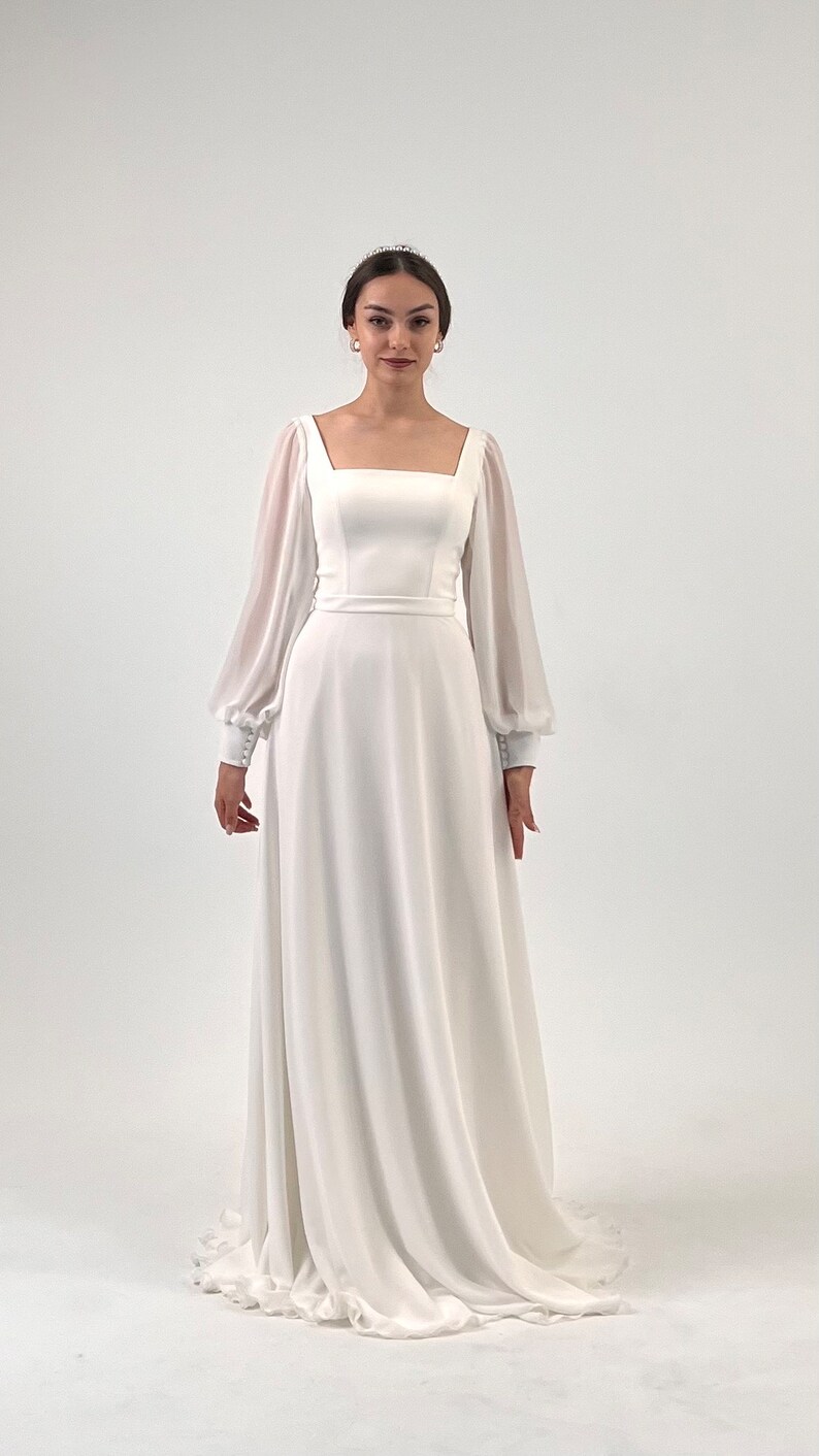Puede incluir: Un vestido de novia blanco con escote cuadrado y mangas largas transparentes. El vestido presenta un corpi&ntilde;o ajustado y una falda fluida. Las mangas tienen pu&ntilde;os abotonados. La modelo lleva una tiara y pendientes.