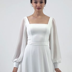 Puede incluir: Un vestido de novia blanco con escote cuadrado y mangas largas y transparentes. El vestido tiene un corpi&ntilde;o ajustado y una falda fluida. Un cintur&oacute;n fino acent&uacute;a la cintura. Las mangas tienen pu&ntilde;os abotonados. Una tiara y pendientes completan el look.