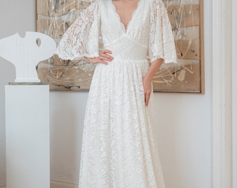 May include: A white lace wedding dress with a V-neckline and bell sleeves. The dress has a fitted bodice and a flowing skirt.