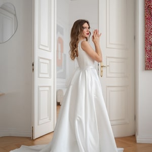 May include: A woman in a white sleeveless wedding dress with a long train stands in a doorway. The dress has a fitted bodice and a flowing skirt.