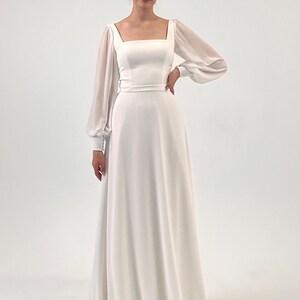 Puede incluir: Vestido de novia blanco con escote cuadrado y mangas largas transparentes. El vestido tiene un corpi&ntilde;o ajustado y una falda en forma de A. Un cintur&oacute;n fino acent&uacute;a la cintura. La novia lleva una tiara y pendientes.