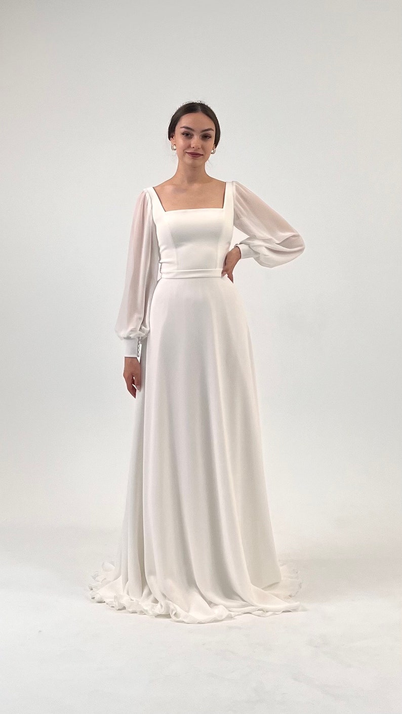 Puede incluir: Vestido de novia blanco con escote cuadrado y mangas largas transparentes. El vestido tiene un corpi&ntilde;o ajustado y una falda en forma de A. Un cintur&oacute;n fino acent&uacute;a la cintura. La novia lleva una tiara y pendientes.