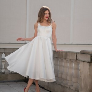 May include: A woman in a white sleeveless dress with a full skirt stands on a balcony. She is wearing red high heels and has her hair down.
