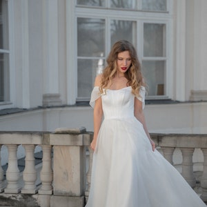 May include: A woman in a white off-the-shoulder wedding dress with a flowing skirt stands on a balcony with a white railing.
