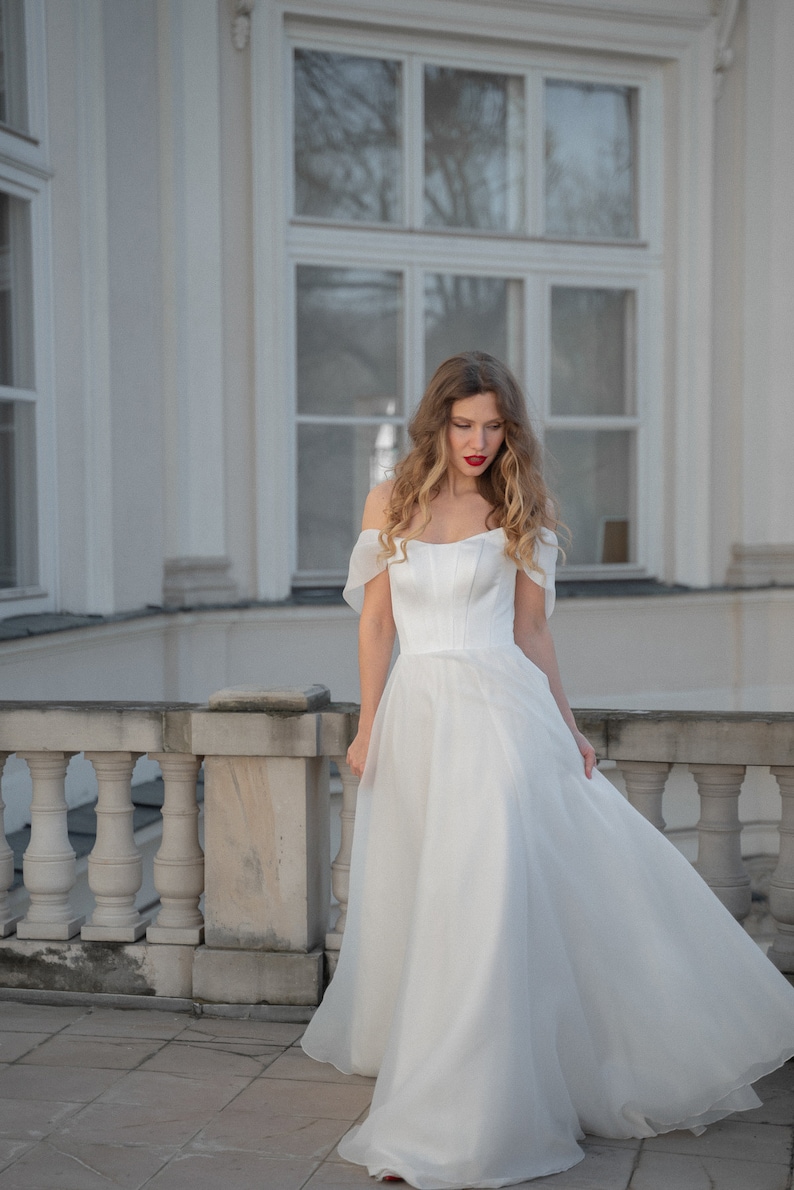 May include: A woman in a white off-the-shoulder wedding dress with a flowing skirt stands on a balcony with a white railing.