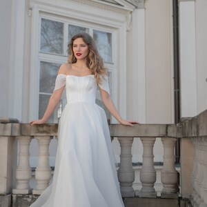 May include: A woman in a white off-the-shoulder wedding dress with a fitted bodice and a flowing skirt stands on a balcony with a stone railing.