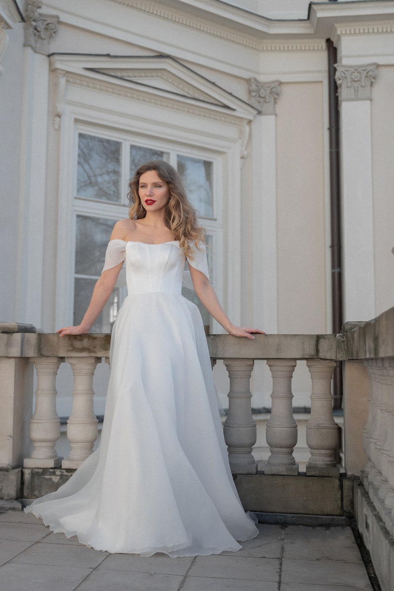 May include: A woman in a white off-the-shoulder wedding dress with a fitted bodice and a flowing skirt stands on a balcony with a stone railing.