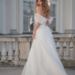 May include: A woman in a white off-the-shoulder wedding dress with a flowing skirt stands on a stone patio in front of a white building.