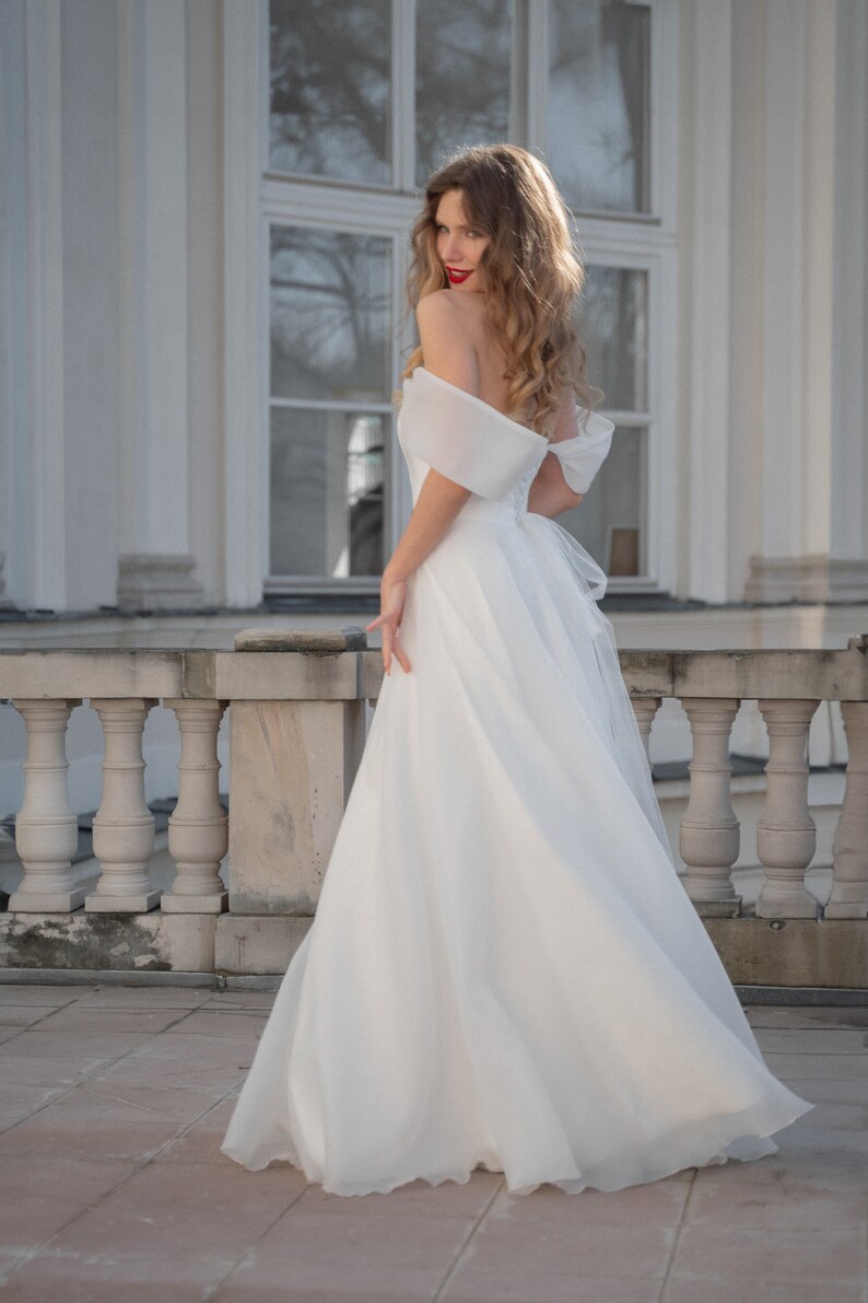 May include: A woman in a white off-the-shoulder wedding dress with a flowing skirt stands on a stone patio in front of a white building.