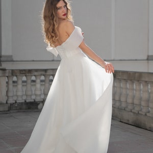 May include: A woman wearing a white off-the-shoulder wedding dress with a flowing skirt. She is standing on a tiled patio with a white railing in the background.