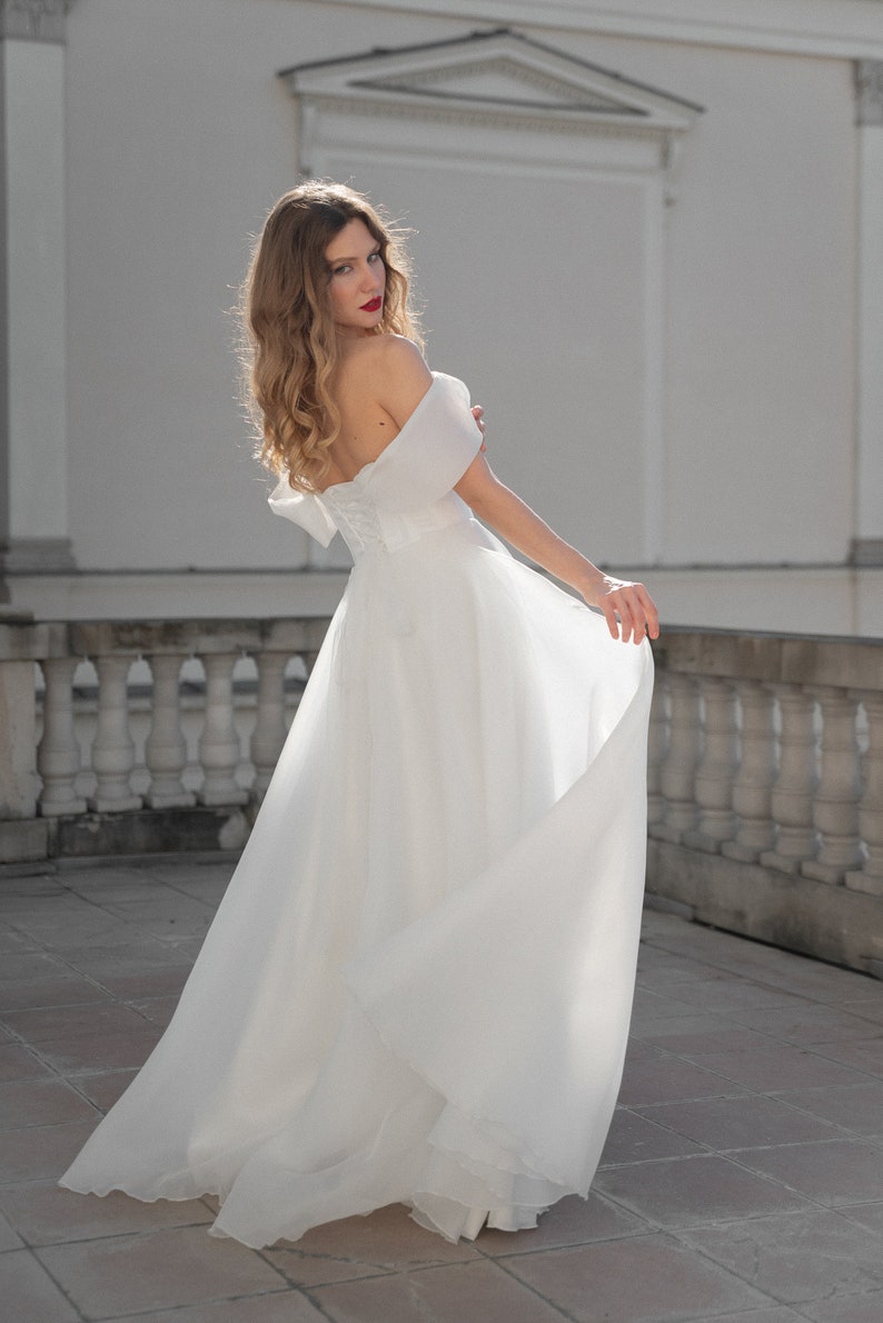 May include: A woman wearing a white off-the-shoulder wedding dress with a flowing skirt. She is standing on a tiled patio with a white railing in the background.