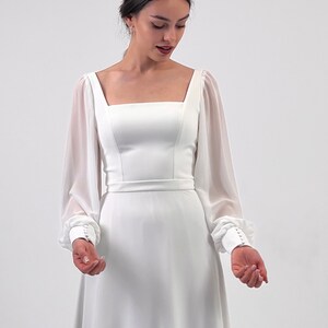 Puede incluir: Vestido de novia blanco con escote cuadrado y mangas largas transparentes. El vestido presenta un corpi&ntilde;o ajustado, una cintura definida y una falda fluida. Las mangas tienen pu&ntilde;os abotonados. Se lleva una diadema de perlas.