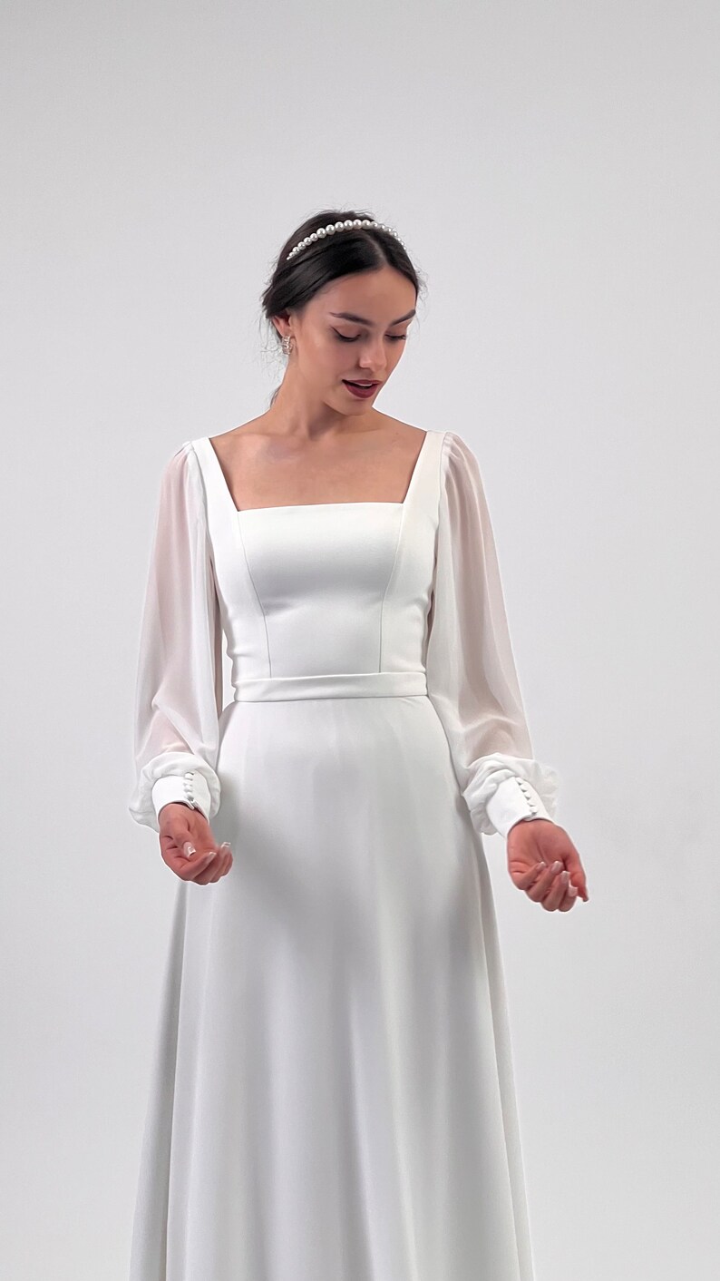 Puede incluir: Vestido de novia blanco con escote cuadrado y mangas largas transparentes. El vestido presenta un corpi&ntilde;o ajustado, una cintura definida y una falda fluida. Las mangas tienen pu&ntilde;os abotonados. Se lleva una diadema de perlas.