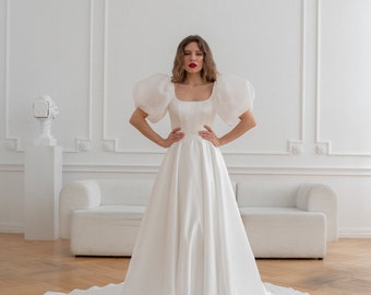 May include: A white wedding dress with a fitted bodice and a full skirt. The dress has puffed sleeves and a square neckline.