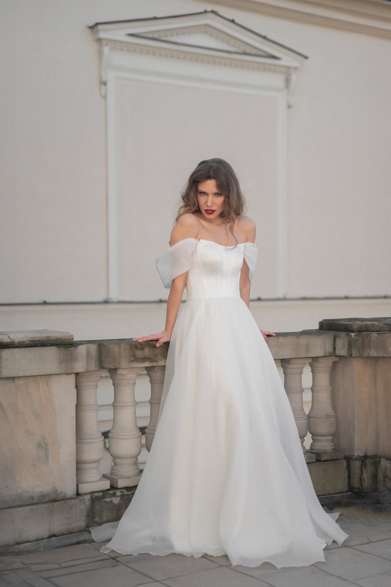 May include: A woman in a white off-the-shoulder wedding dress with a fitted bodice and a flowing skirt. She is standing on a balcony with a white railing.