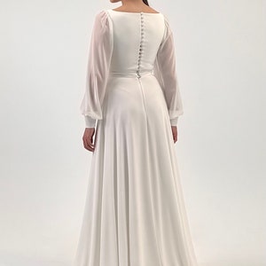 Puede incluir: Un vestido de novia blanco con mangas largas y transparentes y un escote cuadrado. La parte trasera del vestido presenta una fila de botones. La falda llega hasta el suelo. La modelo lleva una diadema y pendientes.